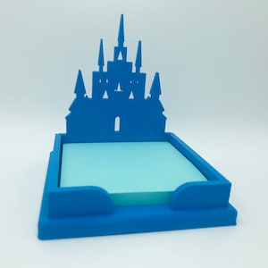 May include: Blue plastic castle-shaped sticky note holder. The holder has a cutout of a castle and a space for a stack of sticky notes.