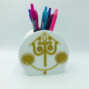 May include: White ceramic pencil holder with a gold design of a stylized tree and two suns. The holder has a round shape and is filled with colorful pens.