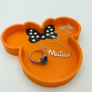 May include: An orange Mickey Mouse-shaped tray with a black and white polka dot bow design. A silver necklace with the name "Melissa" and a ring with a blue gemstone are inside the tray.
