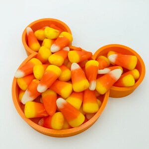 Pu&ograve; includere: Una ciotola arancione a forma di Mickey Mouse piena di caramelle di mais. Le caramelle di mais sono gialle, arancioni e bianche. La ciotola &egrave; di plastica e si trova su una superficie bianca. Una festa per Halloween.