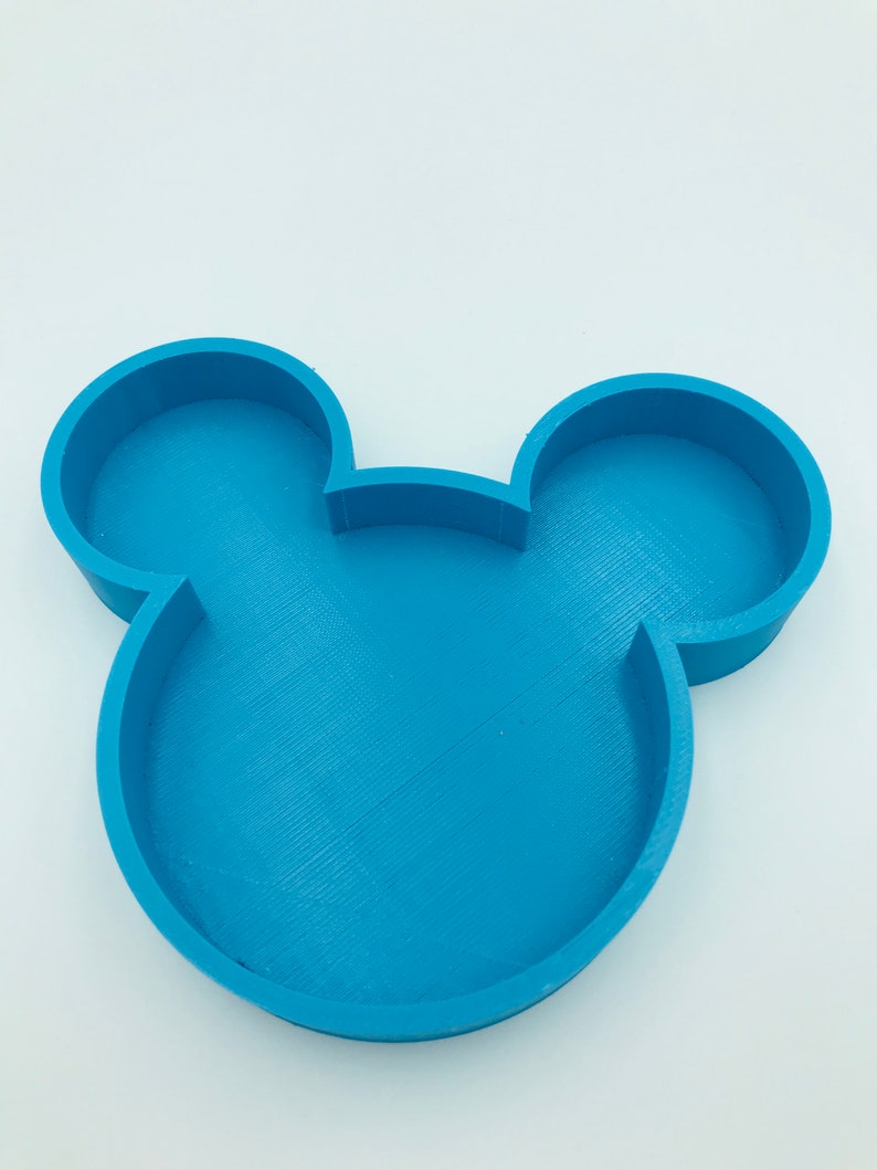 Pu&ograve; includere: Un vassoio blu a forma di Mickey Mouse. Il vassoio &egrave; realizzato in un materiale blu chiaro e presenta le iconiche orecchie del topo. Il vassoio &egrave; progettato con un fondo piatto e bordi rialzati. Lo sfondo &egrave; bianco.
