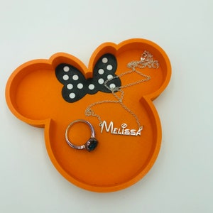 May include: Orange resin jewelry tray in the shape of a mouse head with a black and white polka dot bow. A silver necklace with the name "Melissa" and a ring with a dark stone are inside the tray.