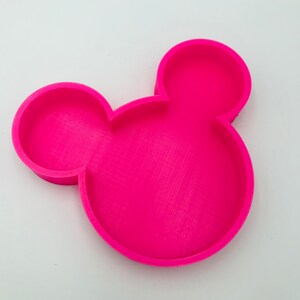 Pu&ograve; includere: Un vassoio rosa acceso a forma di testa di Topolino. Presenta una superficie strutturata, due orecchie circolari e una testa circolare pi&ugrave; grande, con un bordo rialzato. Il vassoio &egrave; realizzato in materiale solido e poggia su uno sfondo bianco.