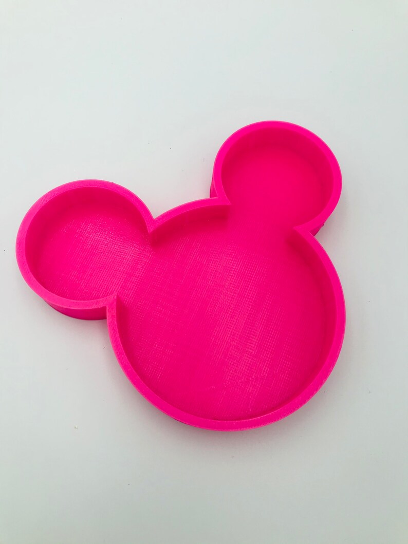 Pu&ograve; includere: Un vassoio rosa acceso a forma di testa di Topolino. Presenta una superficie strutturata, due orecchie circolari e una testa circolare pi&ugrave; grande, con un bordo rialzato. Il vassoio &egrave; realizzato in materiale solido e poggia su uno sfondo bianco.