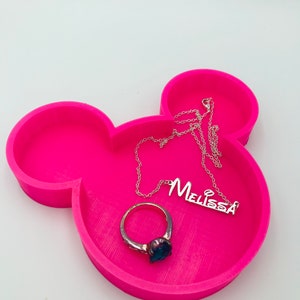 Pu&ograve; includere: Un vassoio rosa acceso a forma di Mickey Mouse con un anello d'argento con una pietra blu e una collana d'argento con il nome "Melissa". Il vassoio &egrave; su una superficie bianca.