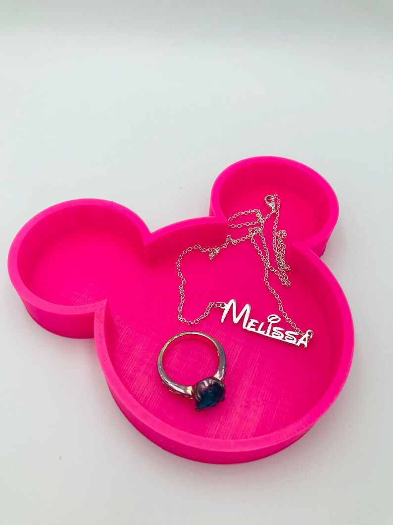 Pu&ograve; includere: Un vassoio rosa acceso a forma di Mickey Mouse con un anello d'argento con una pietra blu e una collana d'argento con il nome "Melissa". Il vassoio &egrave; su una superficie bianca.