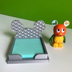 Op de afbeelding: Een zilverkleurige, 3D-geprinte Mickey Mouse-kopvormige kleefnotitiehouder met een lichtblauwe kleefnotitie erin. De houder heeft een geometrisch patroon op de oren en aan de achterkant. Een klein oranje en geel beeldje van een personage met een groen shirt en de tekst "Think Orange Thoughts" is op de achtergrond.