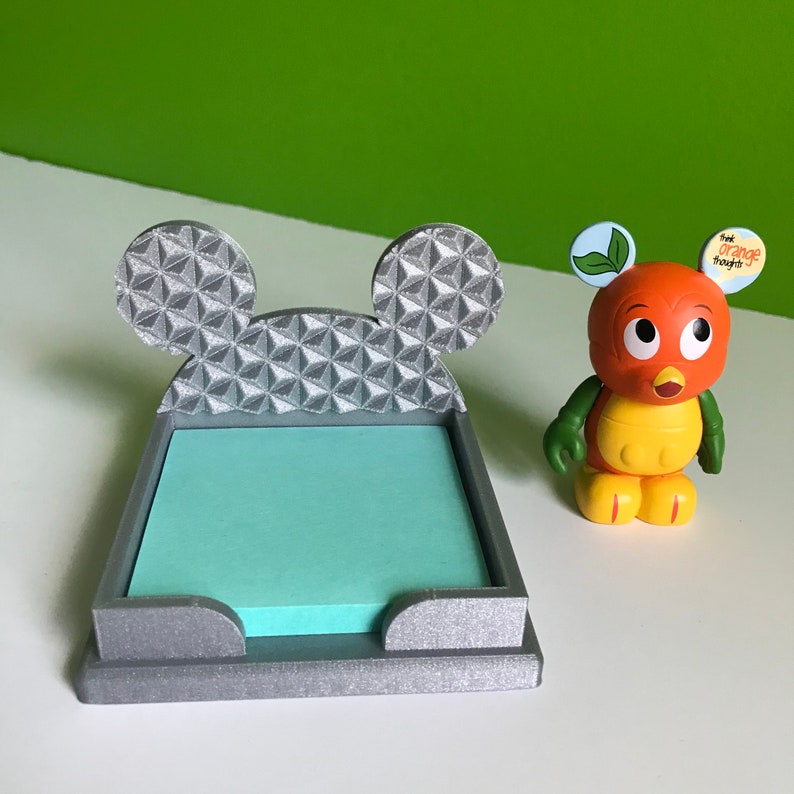 Op de afbeelding: Een zilverkleurige, 3D-geprinte Mickey Mouse-kopvormige kleefnotitiehouder met een lichtblauwe kleefnotitie erin. De houder heeft een geometrisch patroon op de oren en aan de achterkant. Een klein oranje en geel beeldje van een personage met een groen shirt en de tekst "Think Orange Thoughts" is op de achtergrond.