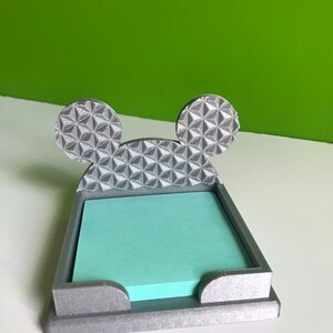 Op de afbeelding: Een zilverkleurige, 3D-geprinte, Mickey Mouse-vormige kleefnotitiehouder. De oren zijn gevormd naar de Spaceship Earth in Epcot.