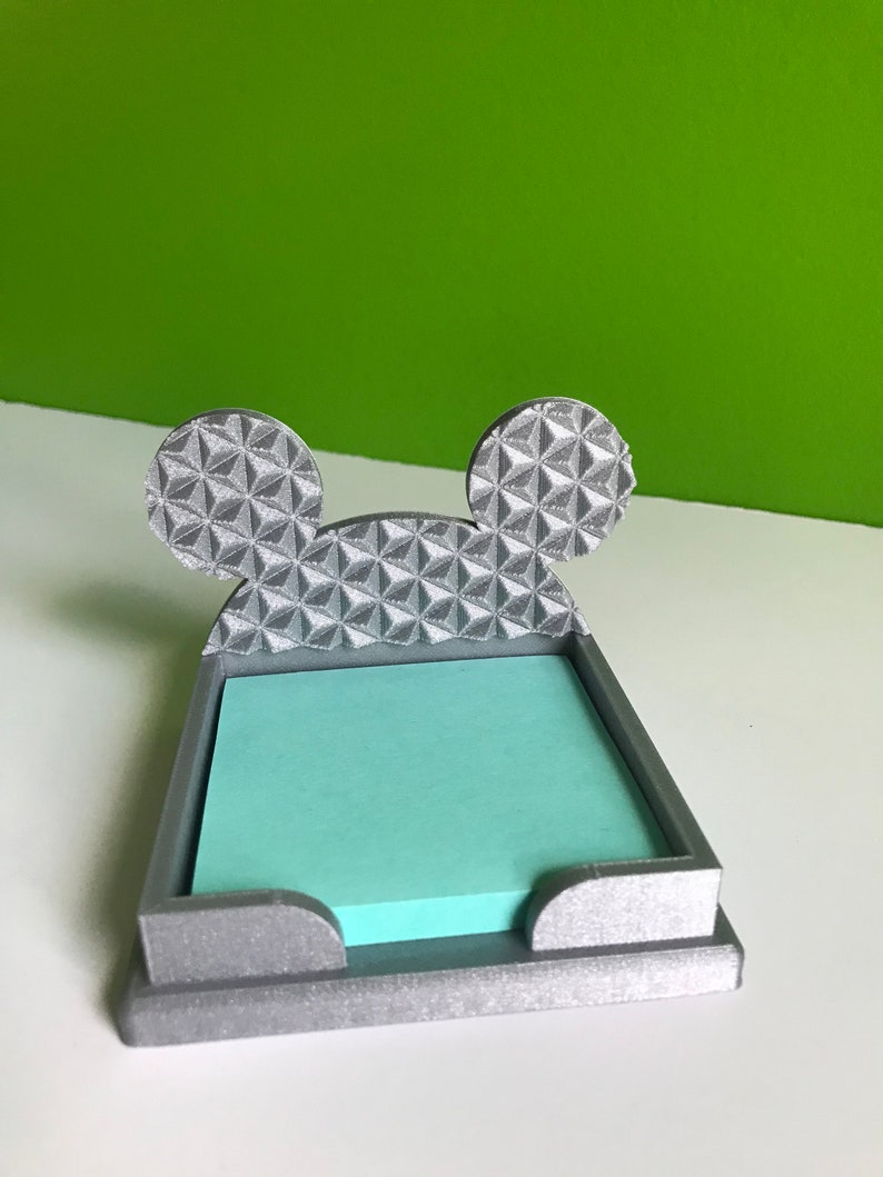 Op de afbeelding: Een zilverkleurige, 3D-geprinte, Mickey Mouse-vormige kleefnotitiehouder. De oren zijn gevormd naar de Spaceship Earth in Epcot.