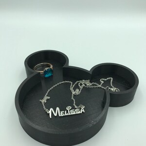 Pu&ograve; includere: Vassoio portagioie nero a forma di Mickey Mouse con una collana d'argento con il nome "Melissa" e un anello con una pietra preziosa blu. Il vassoio &egrave; realizzato in materiale nero opaco.