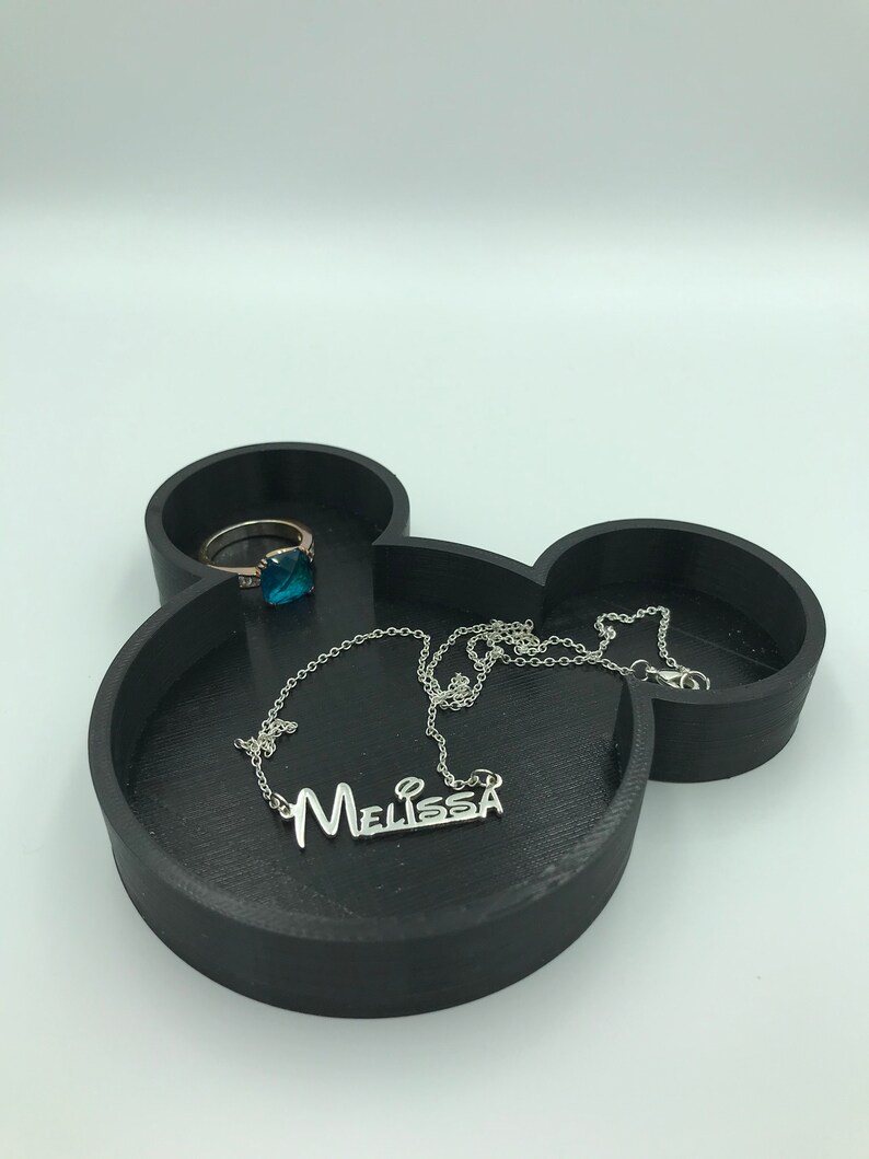 Pu&ograve; includere: Vassoio portagioie nero a forma di Mickey Mouse con una collana d'argento con il nome "Melissa" e un anello con una pietra preziosa blu. Il vassoio &egrave; realizzato in materiale nero opaco.