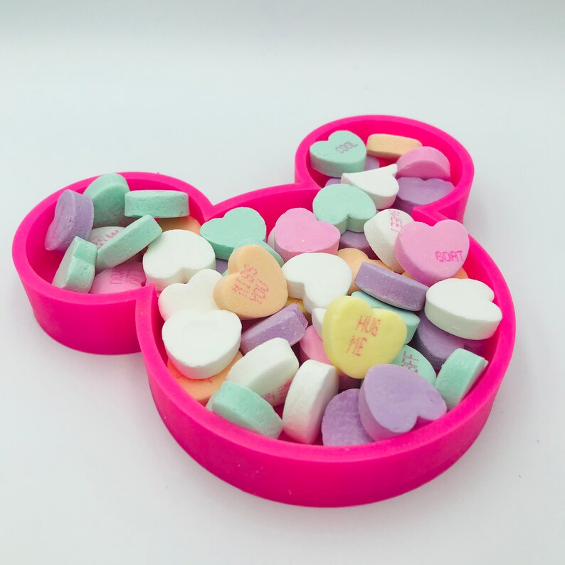 Pu&ograve; includere: Un piatto rosa acceso a forma di Mickey Mouse pieno di caramelle a forma di cuore dai colori pastello. Le caramelle hanno scritte come "HUG ME" e "COOL".