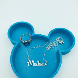Pu&ograve; includere: Un vassoio portagioie blu teal a forma di Mickey Mouse con una collana d'argento con il nome "Melissa" e un anello con una pietra blu. Il vassoio ha tre scomparti circolari.