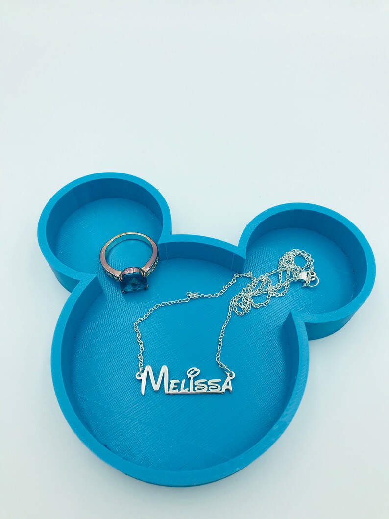 Pu&ograve; includere: Un vassoio portagioie blu teal a forma di Mickey Mouse con una collana d'argento con il nome "Melissa" e un anello con una pietra blu. Il vassoio ha tre scomparti circolari.