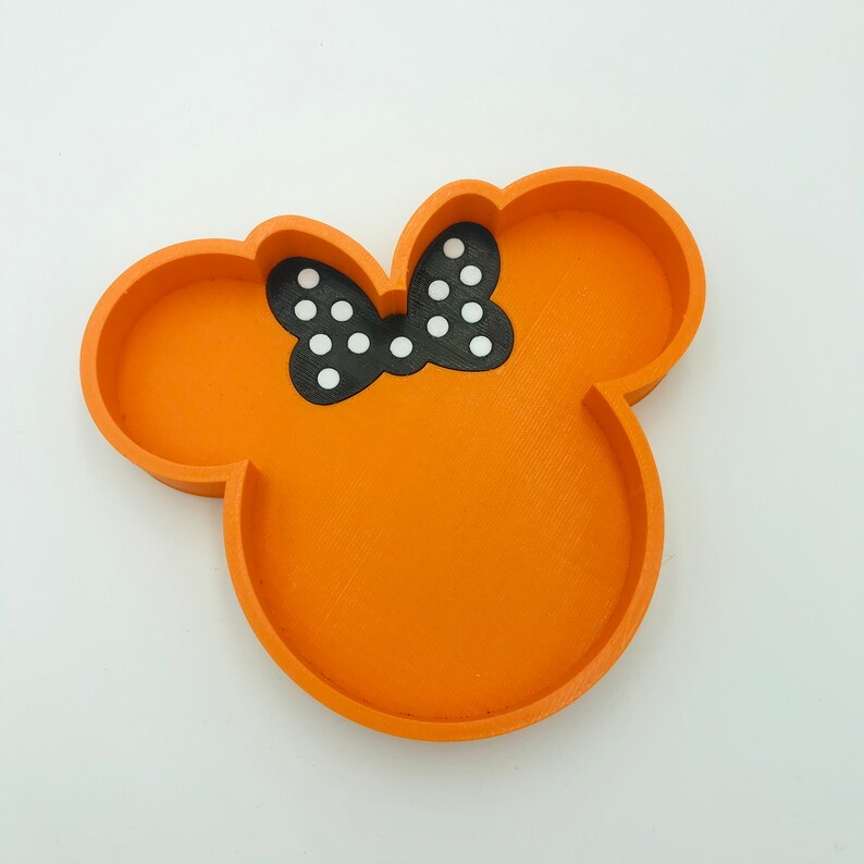 May include: Orange tray in the shape of a mouse head with a black bow featuring white polka dots. The tray has a raised edge and is designed to hold small items. The tray is made of a solid material.