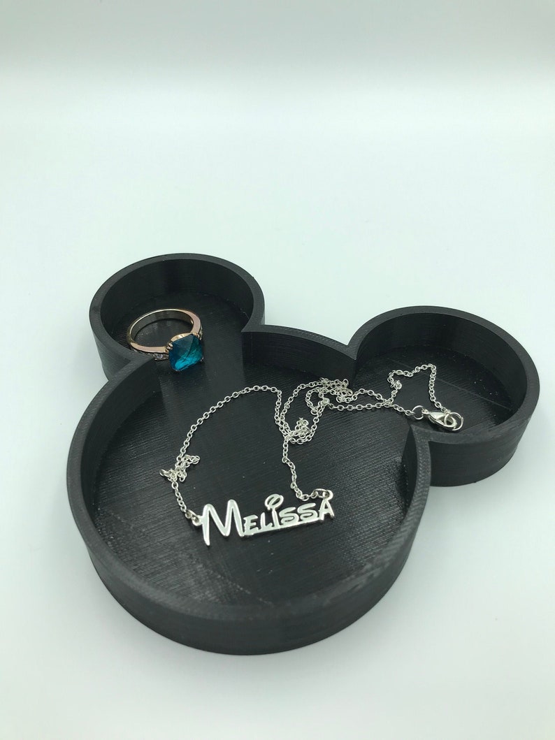 Pu&ograve; includere: Vassoio portagioie nero a forma di Topolino con un anello e una collana. L'anello ha una fascia dorata e una pietra preziosa blu. La collana d'argento riporta il nome "Melissa". Il vassoio &egrave; realizzato in materiale scuro.