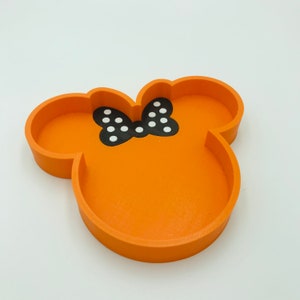 May include: Orange tray in the shape of a mouse head with a black bow featuring white polka dots. The tray is made of wood and has a smooth finish. The design is a playful and decorative item.