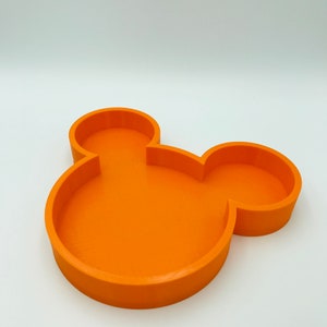 Pu&ograve; includere: Un vassoio arancione a forma di Mickey Mouse. Il vassoio &egrave; di colore pieno e ha una superficie liscia. Il vassoio &egrave; progettato con una base circolare e due forme circolari pi&ugrave; piccole sulla parte superiore, che assomigliano alle orecchie del personaggio iconico. Il vassoio &egrave; probabilmente in plastica.