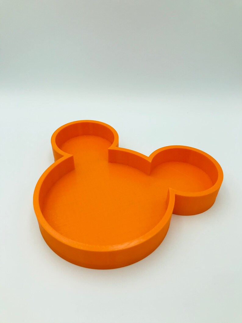 Pu&ograve; includere: Un vassoio arancione a forma di Mickey Mouse. Il vassoio &egrave; di colore pieno e ha una superficie liscia. Il vassoio &egrave; progettato con una base circolare e due forme circolari pi&ugrave; piccole sulla parte superiore, che assomigliano alle orecchie del personaggio iconico. Il vassoio &egrave; probabilmente in plastica.