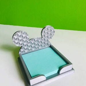 Op de afbeelding: Een zilveren 3D-geprinte notitiehouder in de vorm van Mickey Mouse's hoofd. De oren zijn getextureerd met een geometrisch patroon. Een lichtblauwe kleefnotitie zit in de houder.