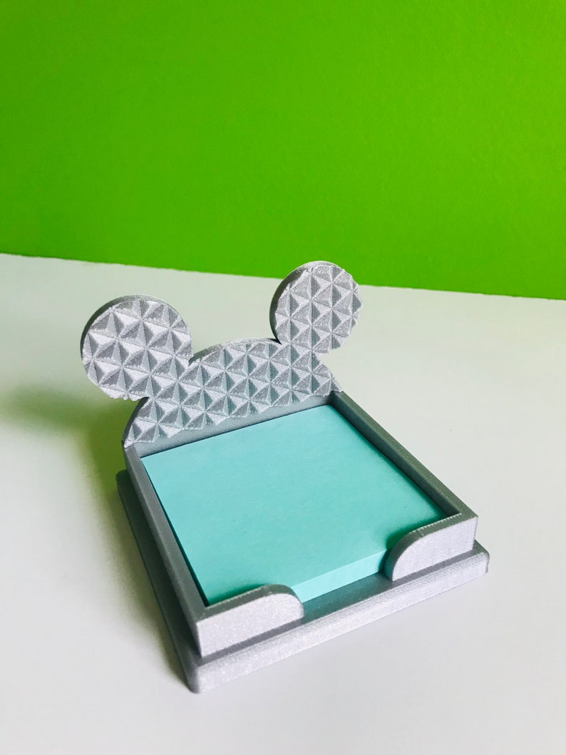 Op de afbeelding: Een zilveren 3D-geprinte notitiehouder in de vorm van Mickey Mouse's hoofd. De oren zijn getextureerd met een geometrisch patroon. Een lichtblauwe kleefnotitie zit in de houder.