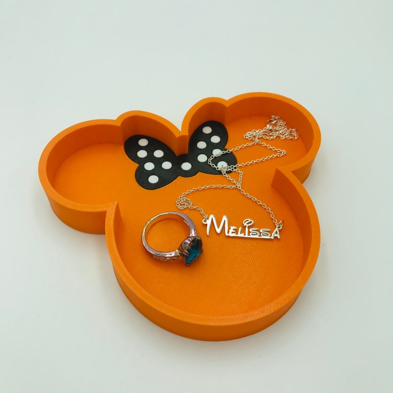 May include: Orange Mickey Mouse-shaped trinket tray with a black and white bow design. A silver necklace with the name "Melissa" and a ring with a blue gemstone are inside the tray. The tray is a decorative item for storing jewelry.