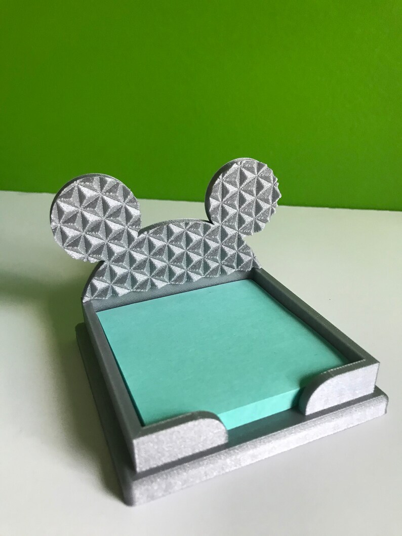 Op de afbeelding: Een grijze 3D-geprinte bureau-organizer in de vorm van Mickey Mouse-oren. De oren zijn getextureerd met een geometrisch patroon. De organizer heeft een gleuf om sticky notes in te bewaren.