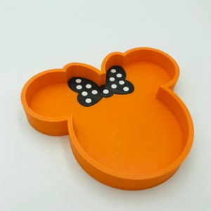 May include: Orange tray in the shape of a cartoon mouse head with a black bow featuring white polka dots. The tray has a raised edge and is designed to hold small items. The tray is made of a solid material.