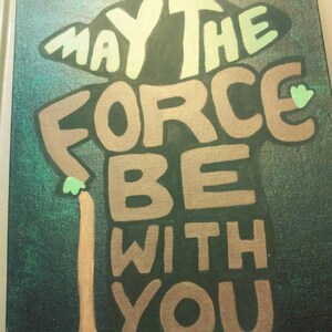 Peut inclure: Une toile peinte à la main en vert et marron avec les mots "Que la Force soit avec toi" écrits dans une police de caractères stylisée. Les mots sont disposés pour former la forme d'un personnage de la franchise Star Wars.