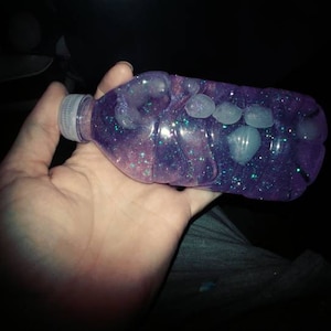 May include: A clear plastic bottle filled with a purple liquid and glitter. The bottle has a white cap and contains small white objects.