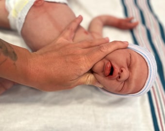 May include: A newborn baby wearing a blue and white striped cap and a diaper. The baby's face is visible, with its mouth open and tongue extended. A hand is gently holding the baby's head.