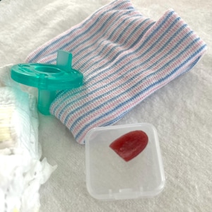 May include: A newborn baby care set featuring a teal pacifier, a striped pink and blue knit cap, and a small plastic case containing a red object. The items are arranged on a white surface, suggesting a hospital or nursery setting.