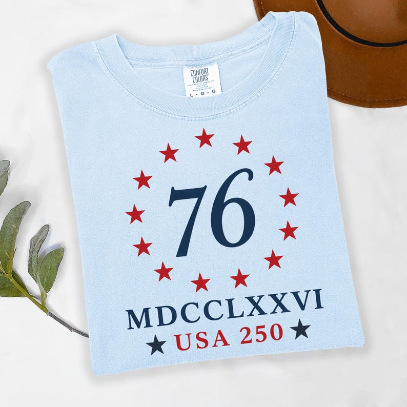 Op de afbeelding: Lichtblauw T-shirt met een ontwerp van rode sterren rond het marineblauwe getal 76. Daaronder de Romeinse cijfers MDCCLXXVI en de tekst USA 250, eveneens in marineblauw. Het shirt is opgevouwen.