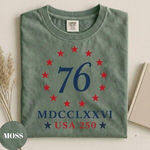 Op de afbeelding: Mosgroen T-shirt met ronde hals. De voorkant toont een marineblauwe "76" omringd door rode sterren, met "MDCCLXXVI" en "USA 250" eronder. Het shirt heeft een vintage, versleten uitstraling.