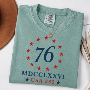 Op de afbeelding: Een saliegroen Comfort Colors T-shirt met het nummer 76 in marineblauw, omcirkeld door rode sterren. Daaronder staan de Romeinse cijfers MDCCLXXVI en de tekst USA 250, eveneens in marineblauw. Een zilveren ketting met hanger ligt op het T-shirt.
