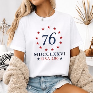 Op de afbeelding: Wit T-shirt met een ontwerp met het nummer "76" in marineblauw, omringd door rode sterren. Daaronder staat "MDCCLXXVI USA 250" in marineblauw met rode sterren. Het shirt heeft een klassieke ronde hals.