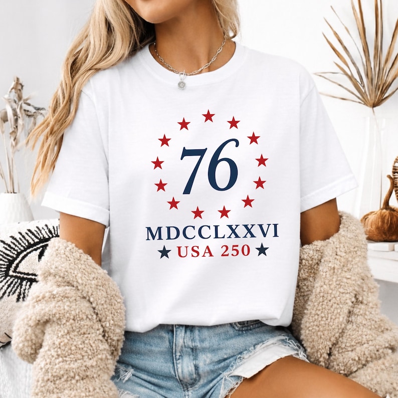 Op de afbeelding: Wit T-shirt met een ontwerp met het nummer "76" in marineblauw, omringd door rode sterren. Daaronder staat "MDCCLXXVI USA 250" in marineblauw met rode sterren. Het shirt heeft een klassieke ronde hals.