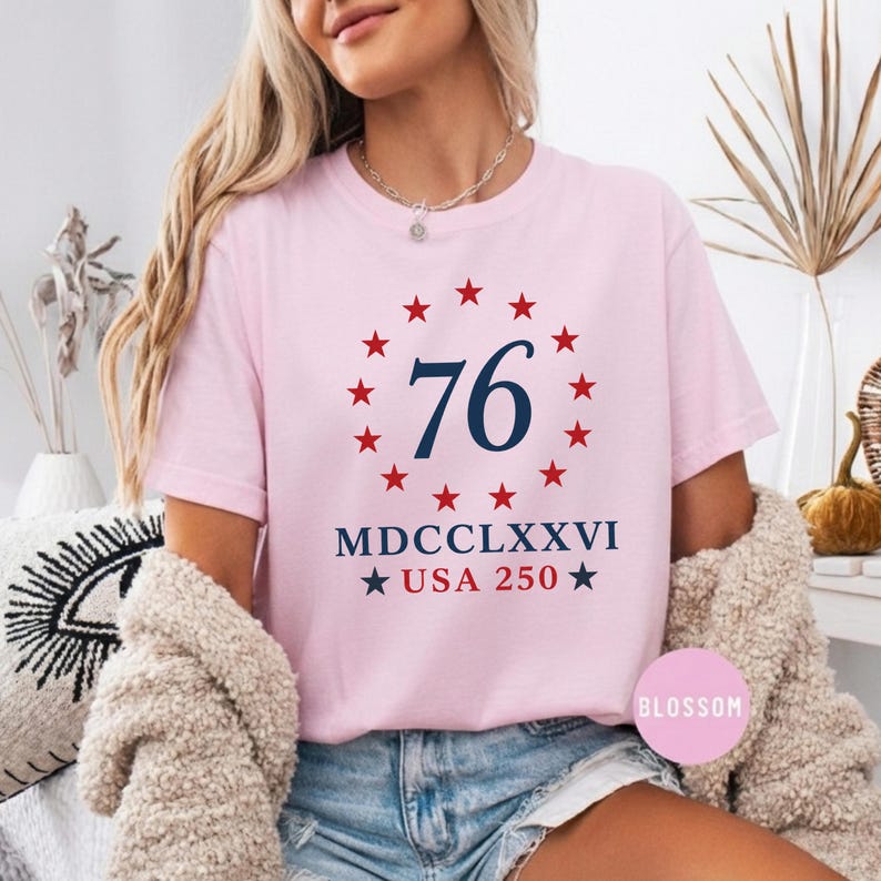 Op de afbeelding: Lichtroze T-shirt met een cirkelvormig ontwerp. Het marineblauwe getal "76" is omringd door rode sterren. Daaronder staan de Romeinse cijfers "MDCCLXXVI" en de tekst "USA 250" met sterren.