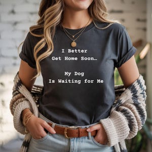Może przedstawiać: Ciemnoszara koszulka z białym napisem "I Better Get Home Soon... My Dog Is Waiting for Me". Koszulka jest połączona z niebieskimi dżinsami i brązowym paskiem. Sweter we wzory jest zarzucony na ramiona.