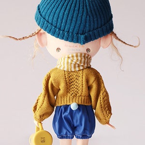 May include: A doll wearing a teal knit hat, mustard yellow sweater, blue shorts, and yellow socks with teal boots. The doll is holding a yellow watering can. The text on the image says "missyodoll" and "Knit Hollow Pattern Cardigan".