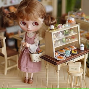 Puede incluir: Una muñeca en miniatura con un vestido rosa está de pie cerca de una pequeña vitrina de panadería llena de pequeños pasteles. La escena incluye una pequeña mesa, un taburete con una caja de pan y una planta en maceta, todo en un entorno de casa de muñecas.