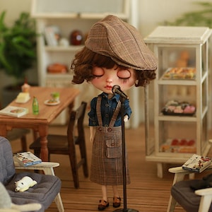 Könnte beinhalten: Eine Puppe, die ein braunes und weißes kariertes Kleid und einen braunen und weißen karierten Hut trägt, hält ein Mikrofon auf einem Ständer. Die Puppe steht in einem Miniaturzimmer mit Möbeln, darunter eine kleine Couch, ein Tisch und ein Regal.
