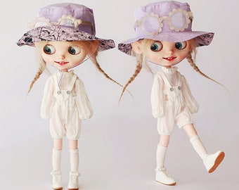 Puede incluir: Dos muñecas con monos blancos y calcetines blancos con sombreros morados. Los sombreros tienen un borde blanco y un diseño de flores blancas. Las muñecas tienen el pelo rubio en trenzas. Las muñecas están de pie sobre un fondo blanco. El texto "Bib Bloomers" es visible en la imagen.