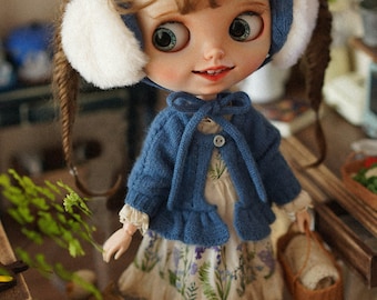May include: A doll wearing a blue knitted cardigan with a white bow, a floral print dress, and white earmuffs. The doll is holding a small wicker basket.