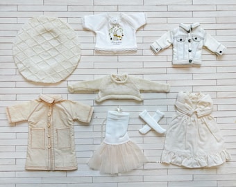 May include: A collection of doll clothing including a white beret, a white t-shirt with a rabbit graphic, a white denim jacket, a beige knit sweater, a beige coat with pockets, a white skirt with a tulle hem, white socks, and a white dress.