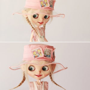 Op de afbeelding: Een pop met een roze vissershoed met een doorzichtige rand en patches met stripfiguren. De pop heeft blonde vlechten, grote ogen en draagt een roze en witte outfit.
