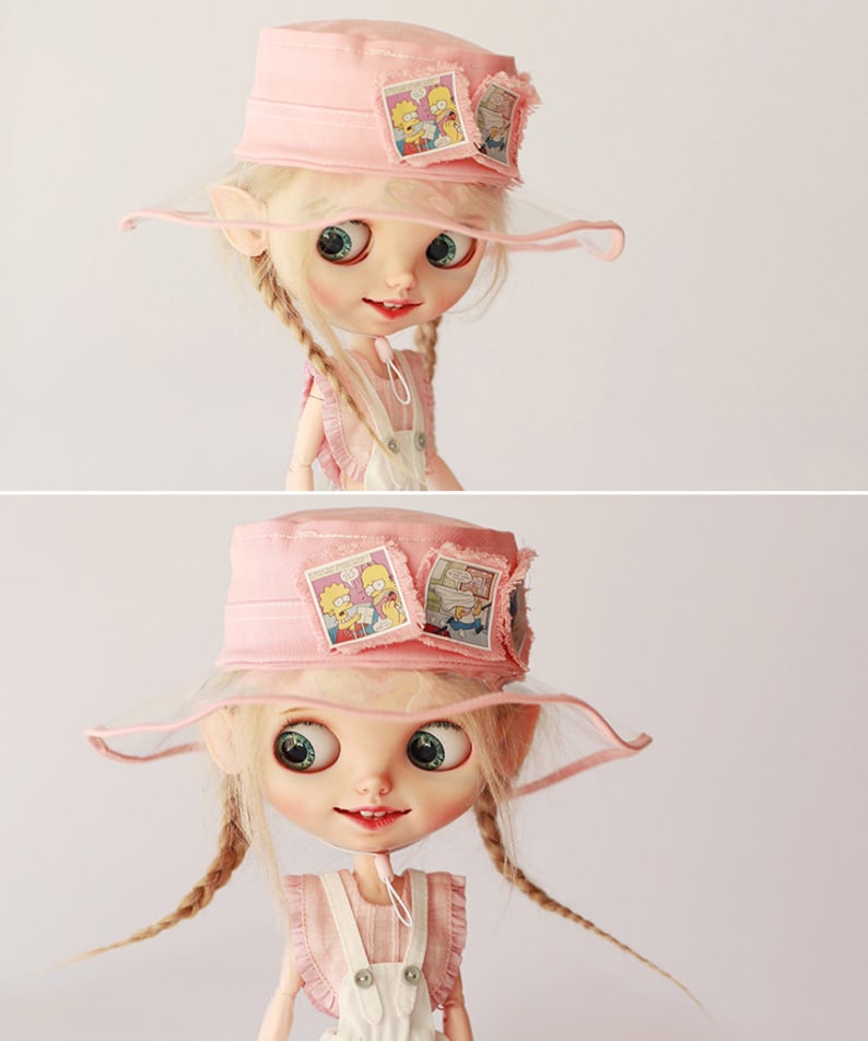 Op de afbeelding: Een pop met een roze vissershoed met een doorzichtige rand en patches met stripfiguren. De pop heeft blonde vlechten, grote ogen en draagt een roze en witte outfit.