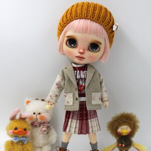 May include: A doll with pink hair and a mustard yellow knit cap, wearing a blazer with floral accents, a red and white shirt, and a plaid skirt. The doll is standing with three plush toys: a duck, a bear, and a lion.