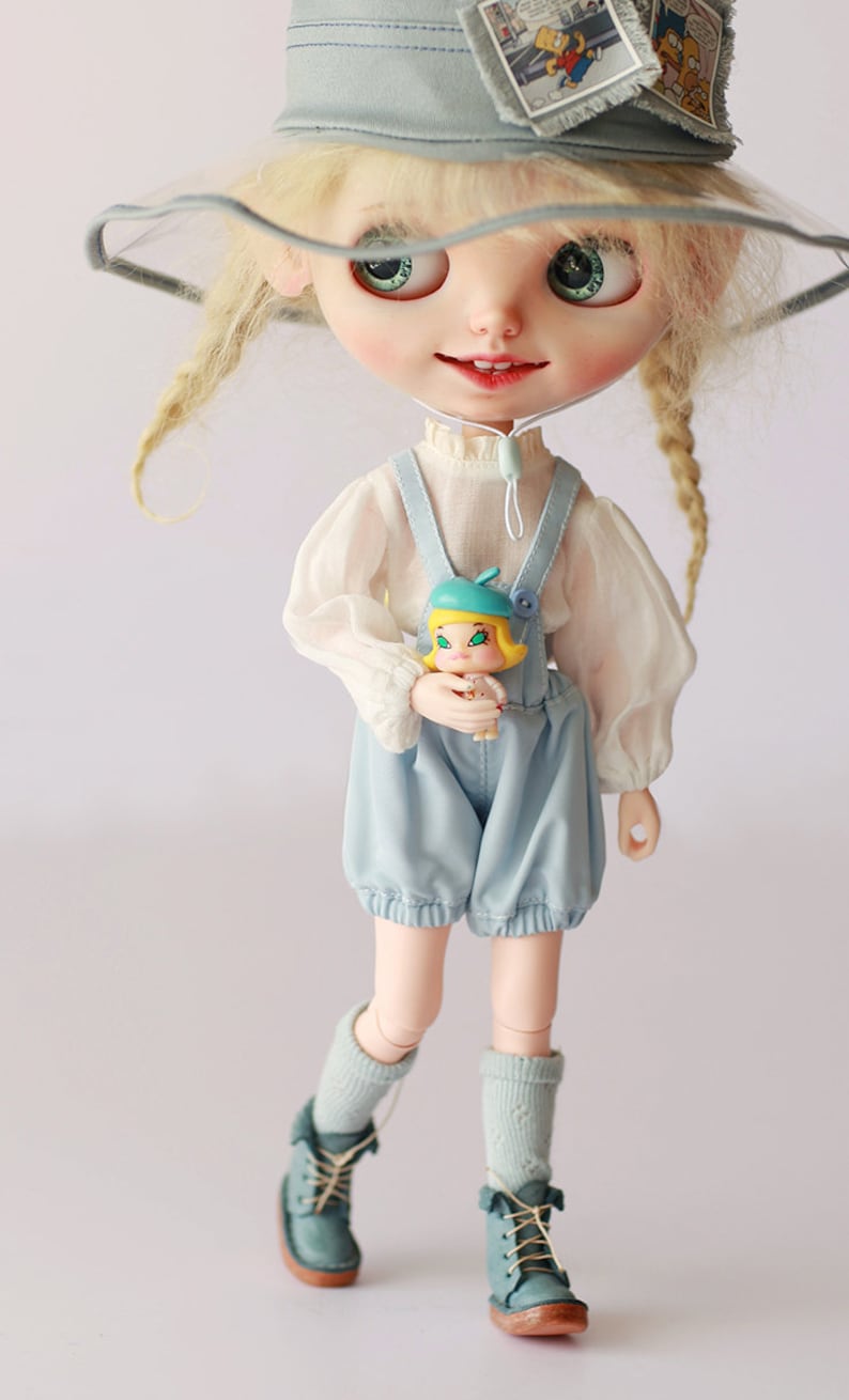Op de afbeelding: Een pop met een lichtblauwe outfit, inclusief een korte broek en bretels, een witte blouse en tealkleurige laarzen. De pop heeft blond haar, groene ogen en een lichtblauwe hoed met stripverhaalpatches. Ze houdt een kleine pop vast.